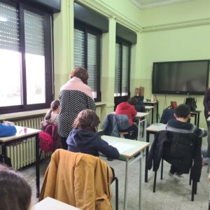 Studenti seduti davanti al banco, in una aula scolastica con pareti tinte di giallo teneue, ripresi di spalle; un insegnante di spalle accanto a uno studente