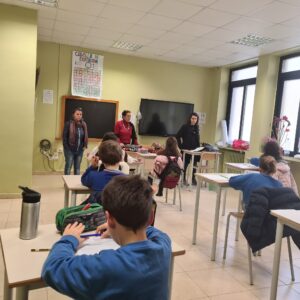 Studenti seduti davanti al banco, in una aula scolastica con pareti tinte di giallo teneue, ripresi di spalle; tre insegnanti in piedi