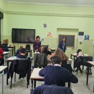 Studenti seduti davanti al banco, in una aula scolastica con pareti tinte di giallo teneue, ripresi di spalle; due insegnanti in piedi 