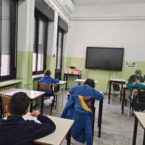 Studenti seduti davanti al banco, in una aula scolastica con pareti tinte di giallo teneue, ripresi di spalle; lungo il margine destro della foto si intravede un'insegnante seduta alla cattedra