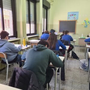 Studenti seduti davanti al banco, in una aula scolastica con pareti tinte di giallo teneue, ripresi di spalle