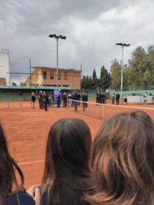 In primo piano studenti di spalle, guardano il campo di tennis, sullo sfondo un gruppo di persone